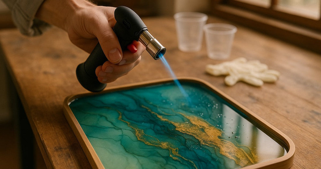 Artist using a butane torch to remove bubbles from a freshly poured epoxy resin tray with teal and gold alcohol ink patterns
