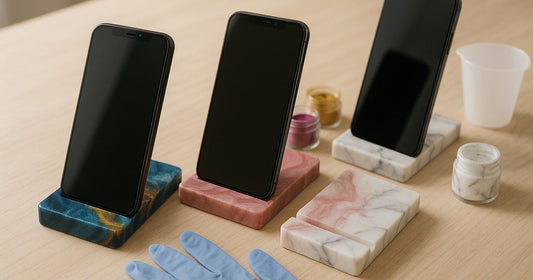Three handmade resin mobile stands in ocean blue, dusty rose, and white marble finishes displayed on a wooden desk with craft materials