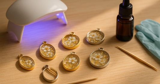 Metal pendant bezels filled with clear UV resin embedding dried flowers and gold foil, curing under a mini UV lamp on a wooden craft table